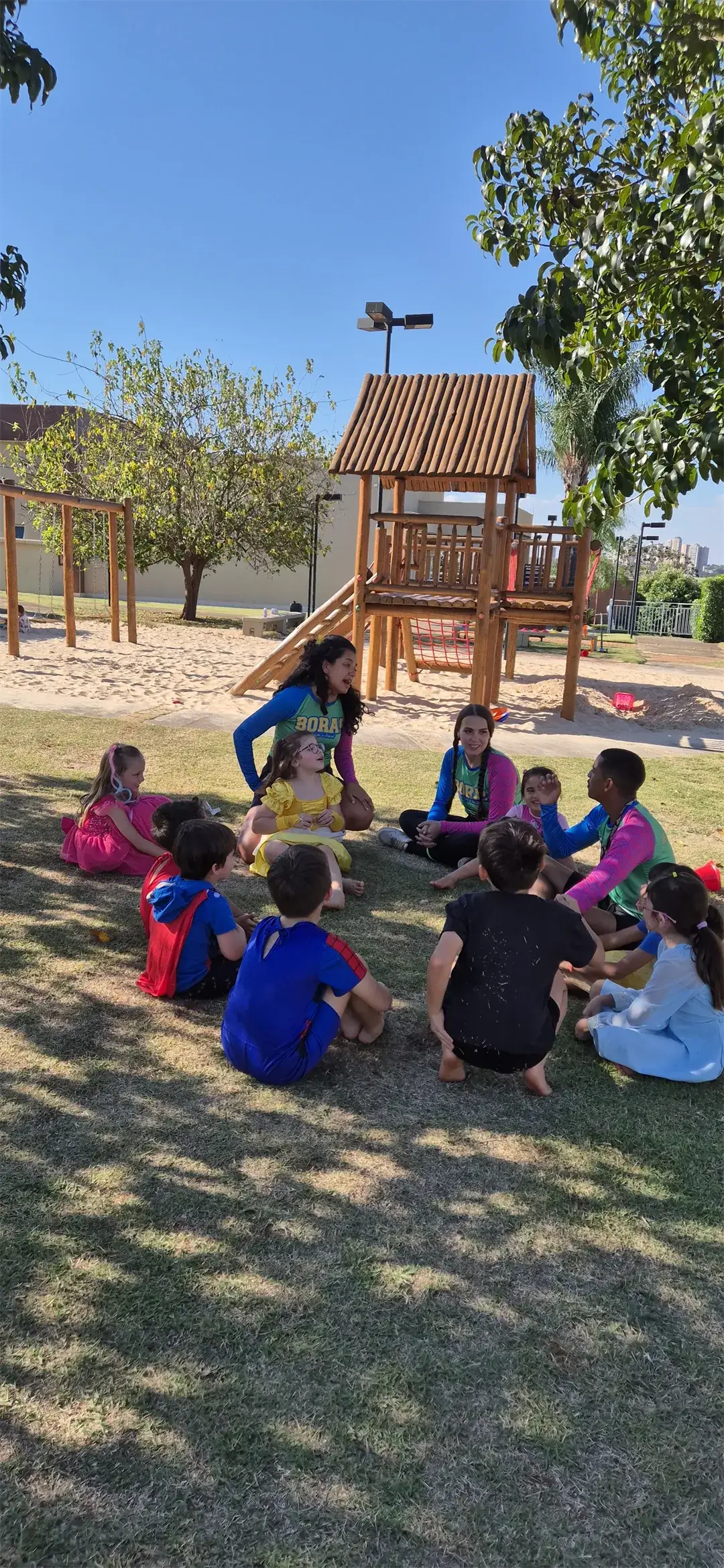 Crianças sentadas em roda com recreadores no gramado, perto de um playground